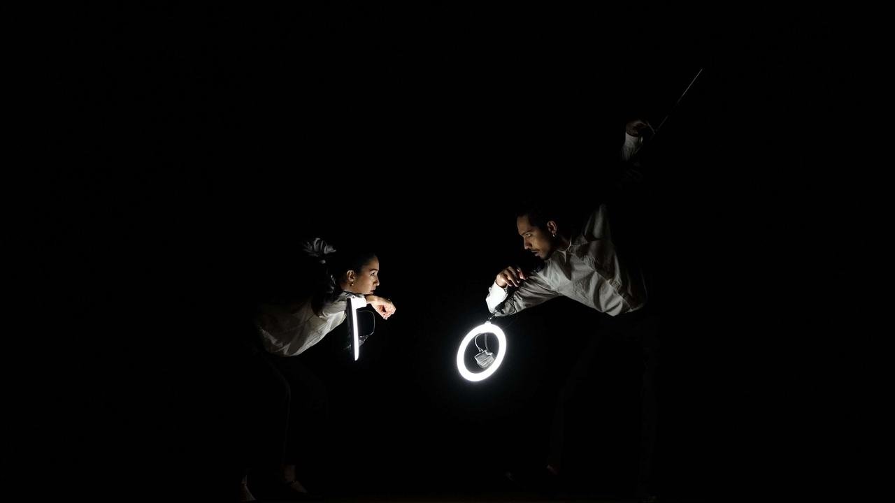 Dois atores em cena no palco escuro, iluminados por um anel de luz